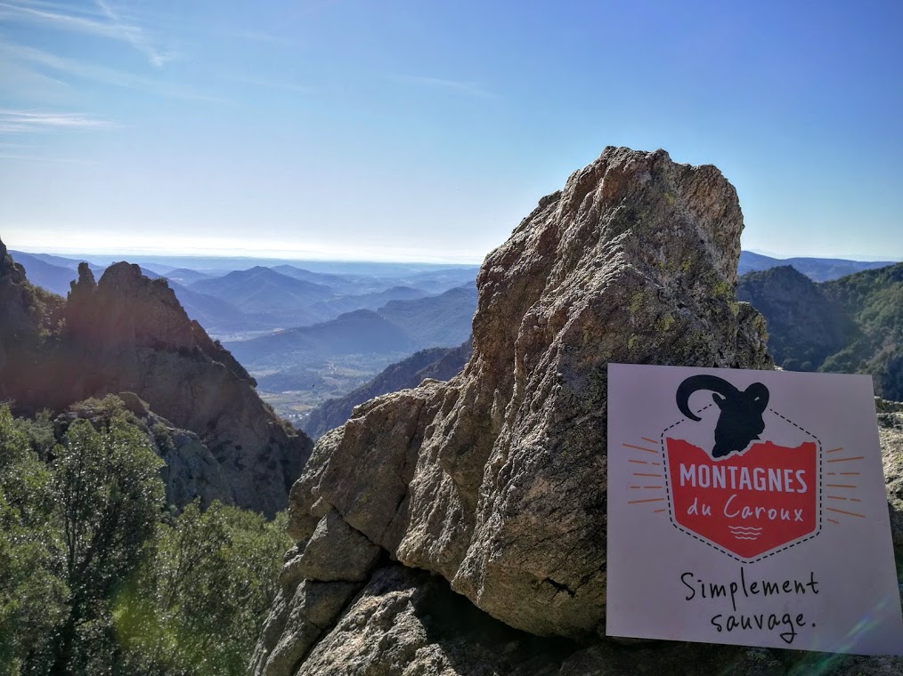 Le logo du pôle de pleine nature "Montagnes du Caroux" exposé sur un sommet du caroux avec en toile de fond le paysage naturel et surplombant depuis les montagnes du caroux